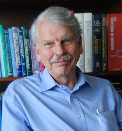 Stephen Hulley, who has white hair and a mustache, smiles in front of a bookcase